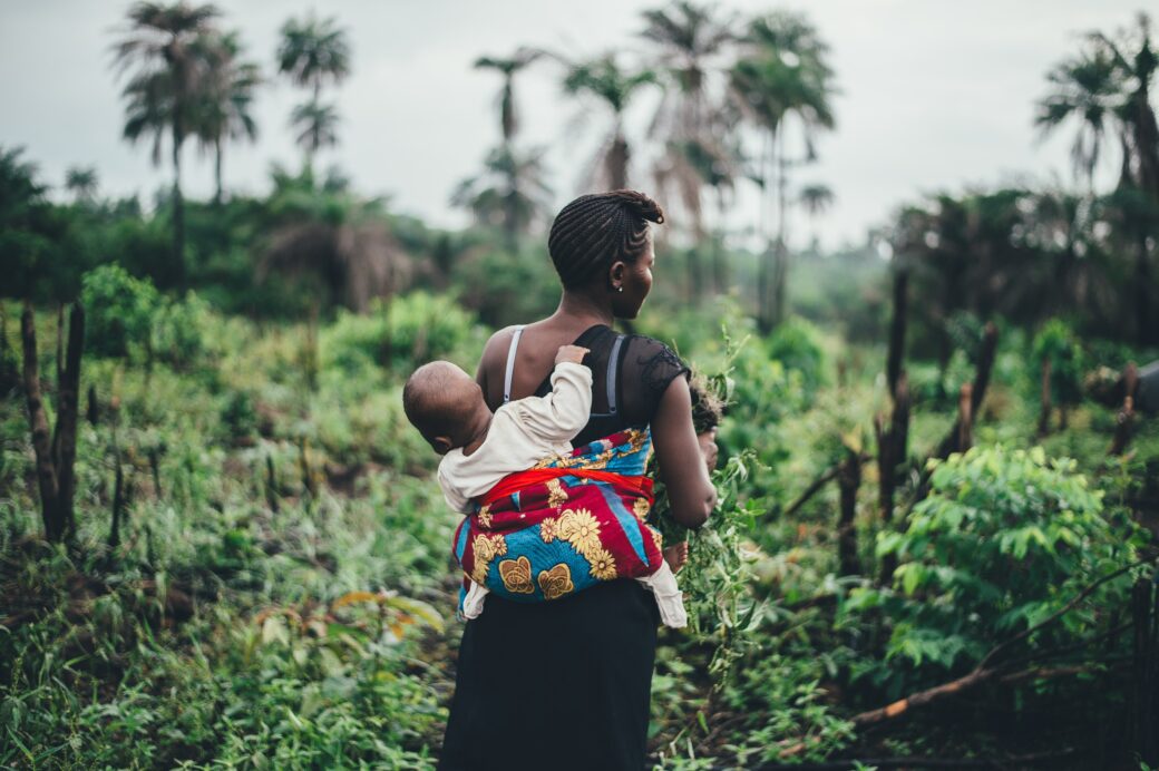 A Black woman in Sierra Leone carrying a child on her back and standing in a forest