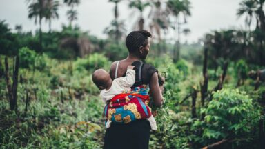 A Black woman in Sierra Leone carrying a child on her back and standing in a forest