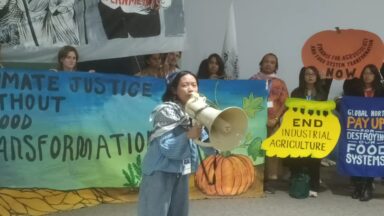 Asian woman speaking into megaphone in front of a protest action at cop29